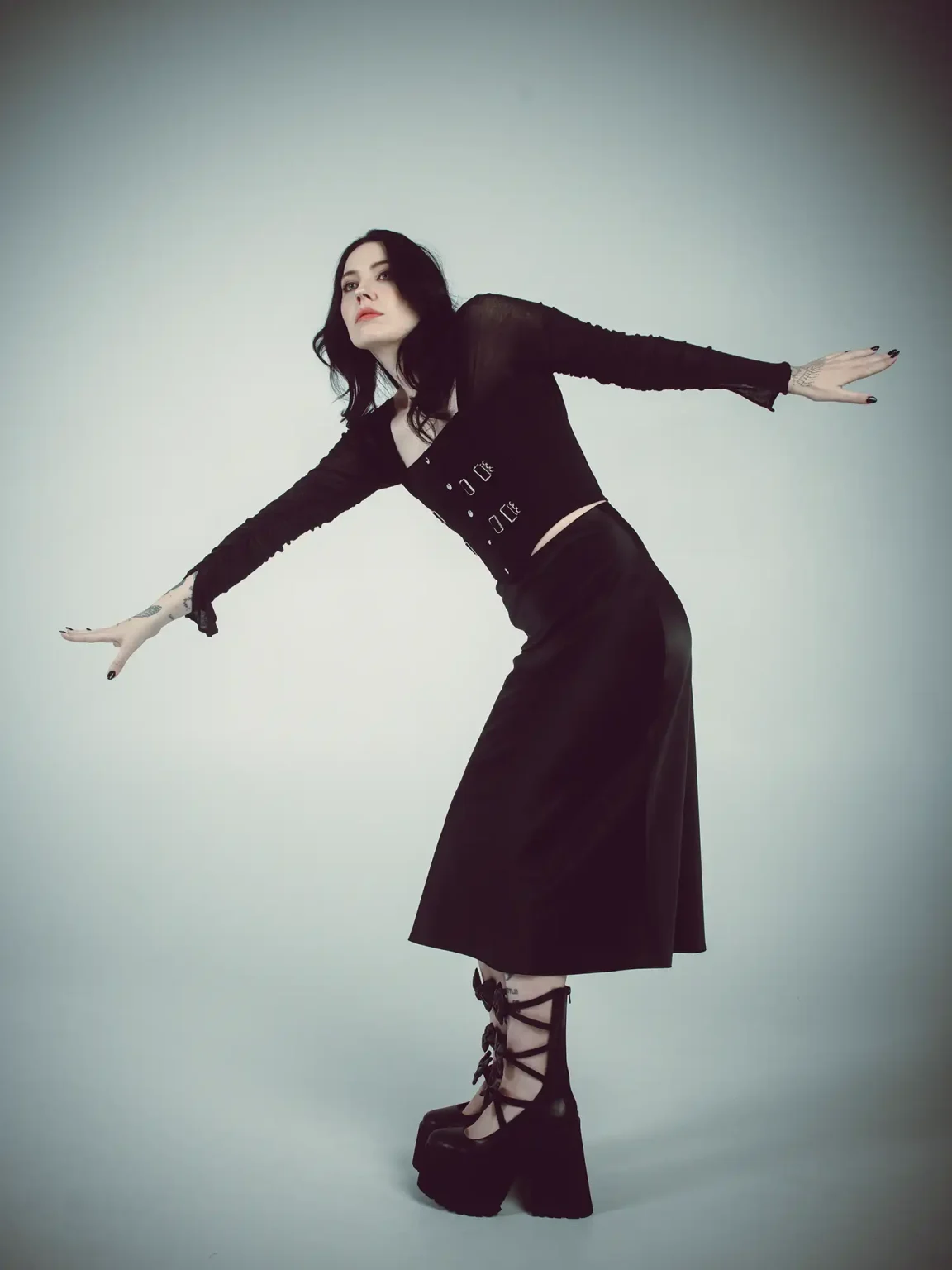 Bishop Briggs poses against a pale grey background in all black — sheer long-sleeve top, midi skirt, and strappy platform boots — arms outstretched, leaning into the frame with effortless energy.