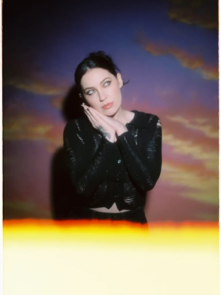 Bishop Briggs gazes upward against a dreamy painted sunset backdrop — warm golds and purples — hands pressed together at her cheek, in a black textured jacket. Shot on film with a warm light leak bleeding across the bottom of the frame.